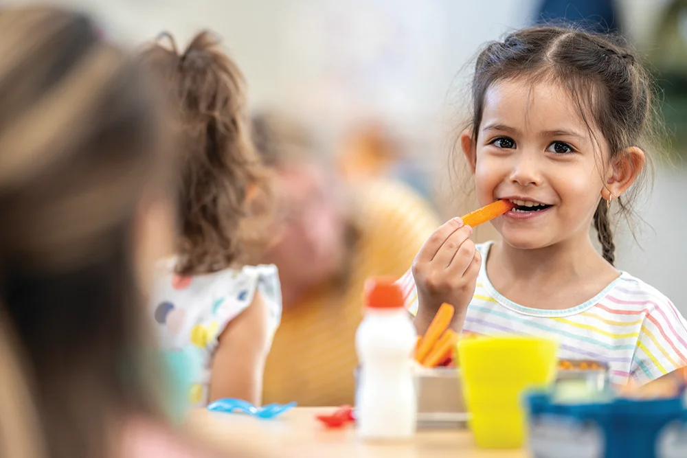 Girl eating carrot at lunch table smiling
